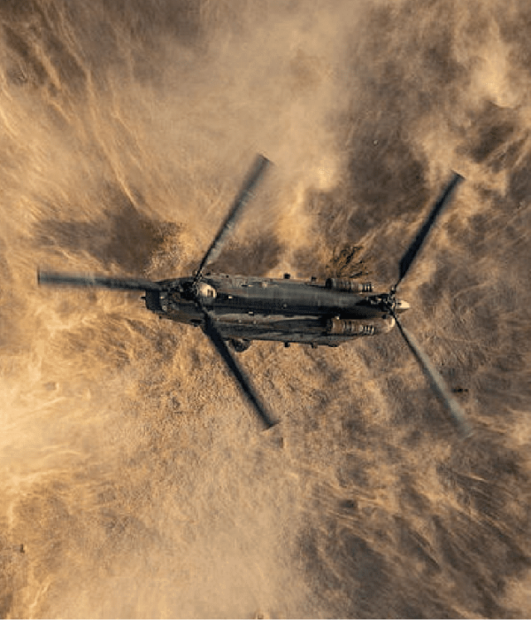 Image-1 A military helicopter flies over a sandy landscape, kicking up a cloud of dust beneath its spinning rotors.
