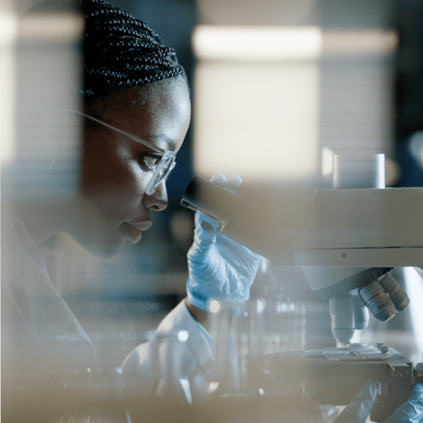 Lab-Tech_1@2x A scientist wearing gloves and glasses looks into a microscope in a laboratory setting.