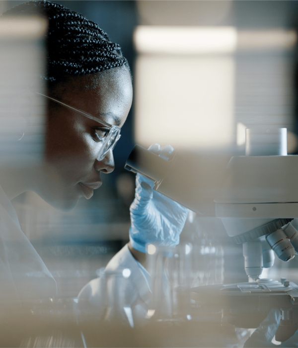 Lab-Tech_1@2x A scientist wearing gloves and glasses looks into a microscope in a laboratory setting.