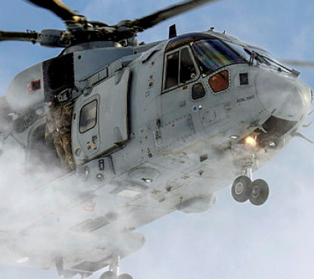 Grey military helicopter hovers low in snowy conditions with open door and soldiers visible inside.