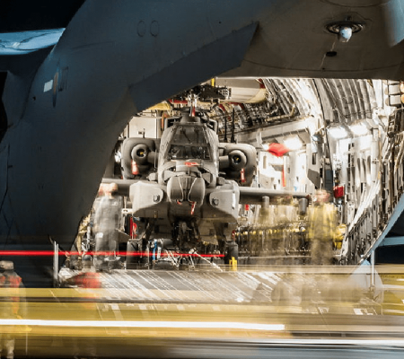 A military helicopter is loaded inside a large cargo aircraft, with crew members standing nearby.