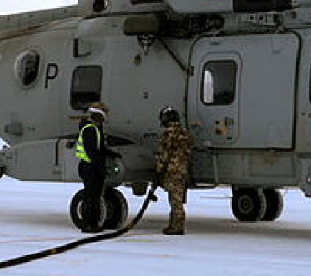 Two people refuel a military helicopter on a snowy surface.