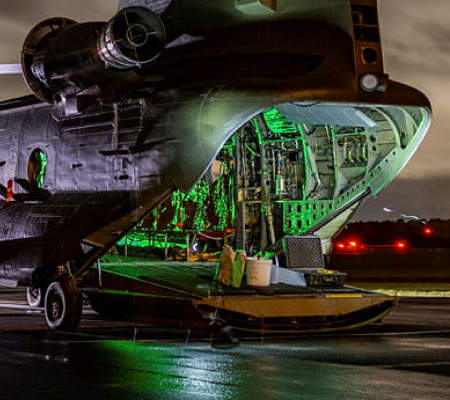 A military cargo helicopter with its rear ramp open, illuminated by green light at night.