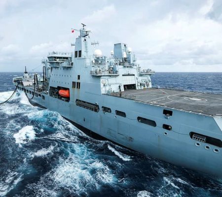 A large gray naval ship sails through choppy ocean waters under a cloudy sky.