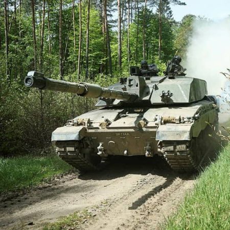 A military tank moving on a dirt road through a forest, with smoke visible behind it.