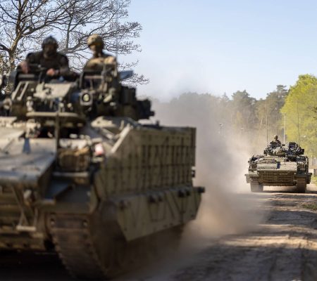 Warrior - DDC-OFFICIAL-20230507-180-004 Two military armored vehicles drive on a dirt road, raising dust, with trees in the background.